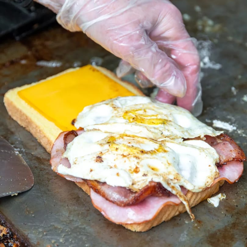 A gloved hand assembling a breakfast sandwich with cheese, ham, bacon, and fried eggs on toast.