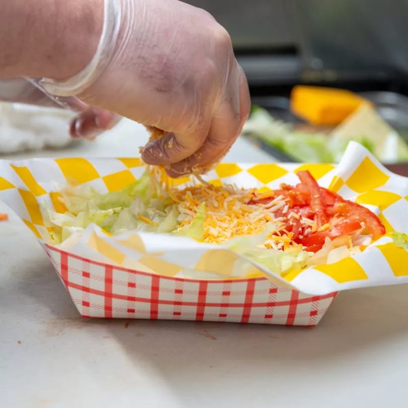 A gloved hand sprinkles shredded cheese onto lettuce and diced tomatoes in a checkered paper tray.
